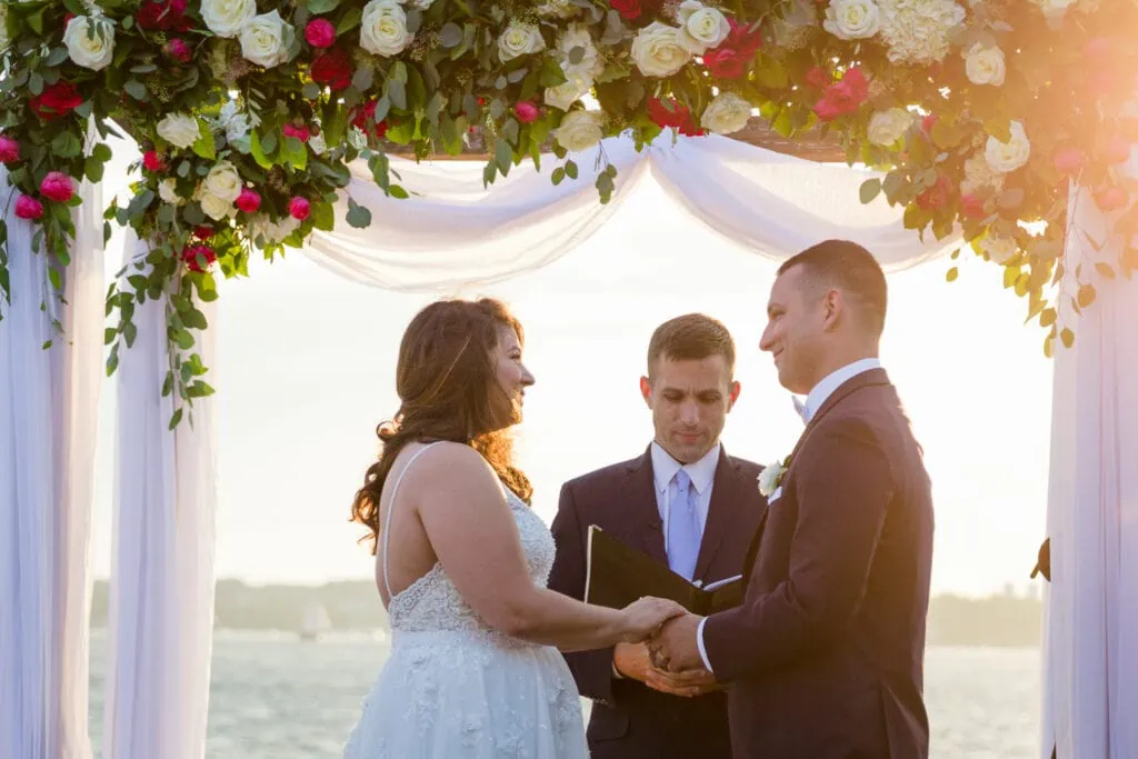 A bride and groom look at each other holding hands at their wedding ceremony with florals and sunset light surrounding them