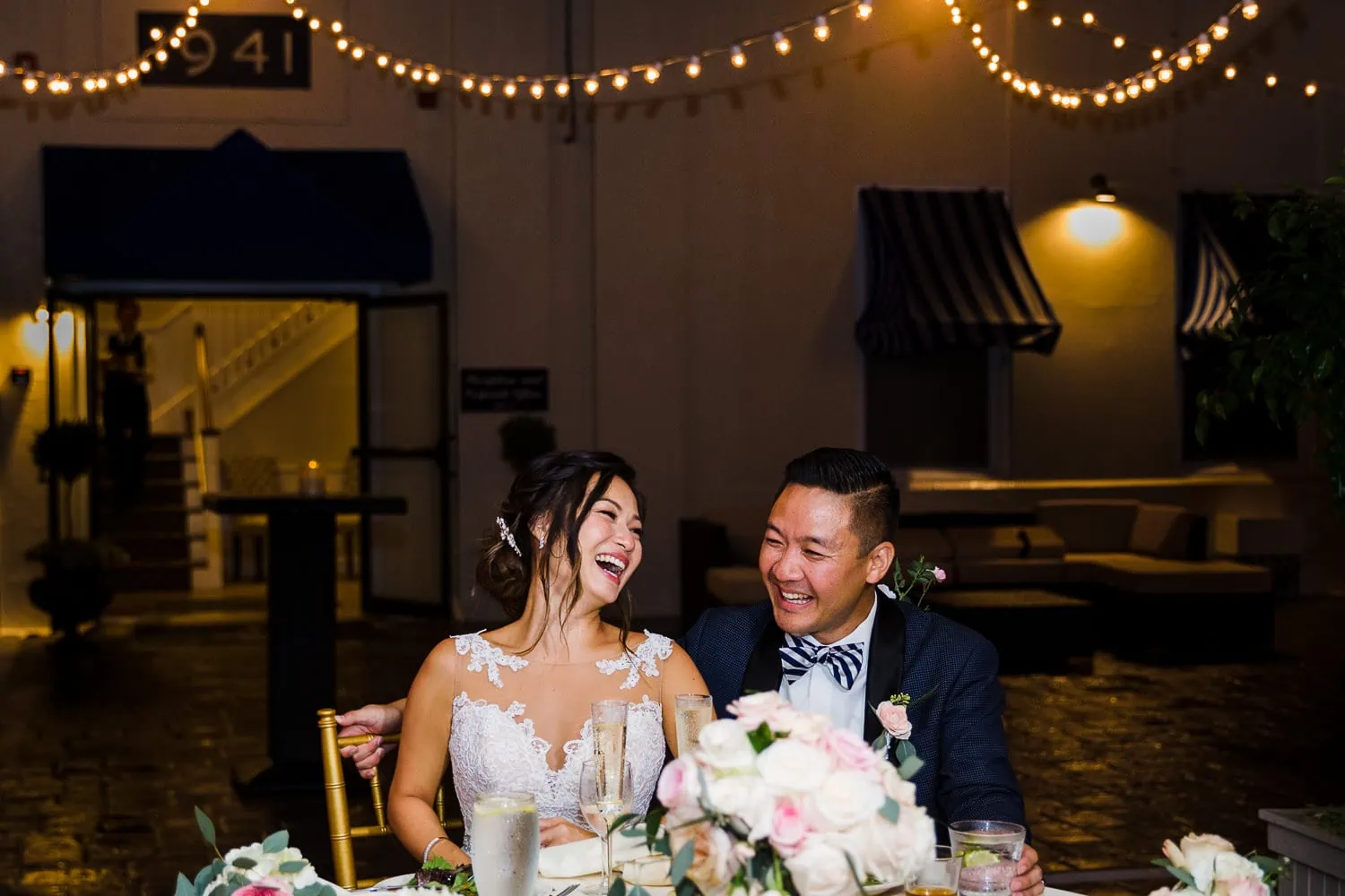 A bride and groom laughing at a sweetheart table with string lights above them