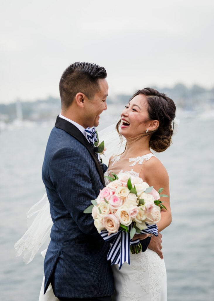 A bride and groom embrace and laugh with the ocean behind them during their regatta place wedding photo session