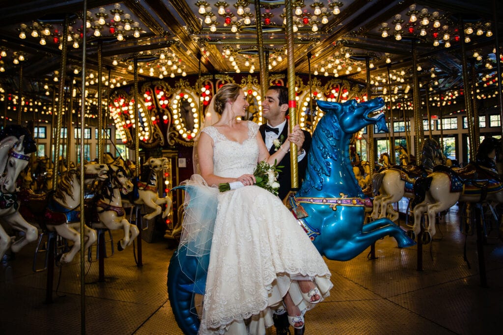 A bride sitting on a carousel hours kisses her groom during wedding photos at roger williams park in the carousel