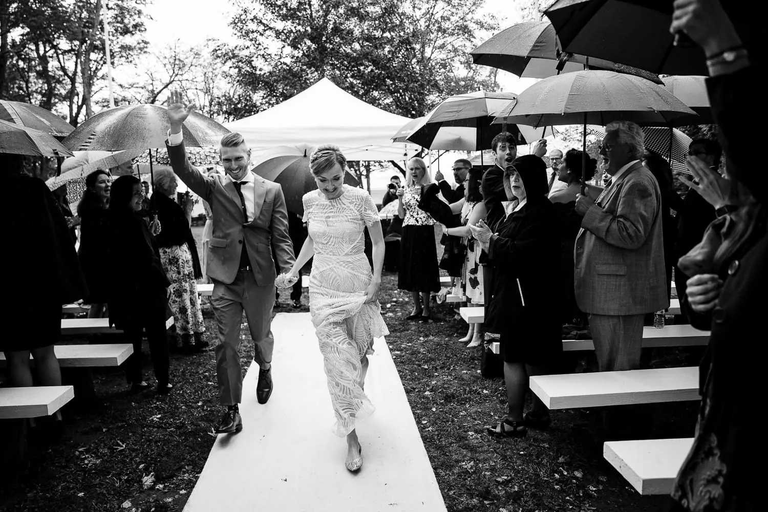 A bride and groom run hand in hand through the rain away from their wedding ceremony as guests under umbrellas look on