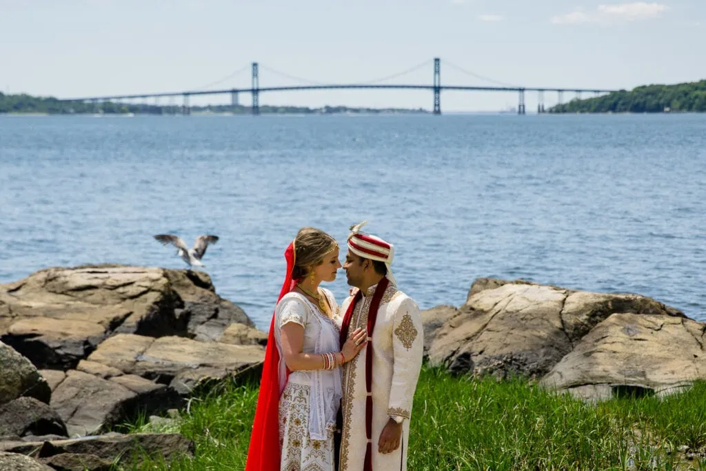 A bride and groom in traditional indian wedding attire standing in the grass with a seagull, ocean and the mount hope bridge behind them