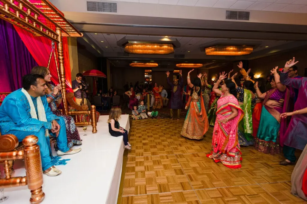 A couple sits on a stage and watches the aunties performing a dance for their indian fusion wedding Sangeeet