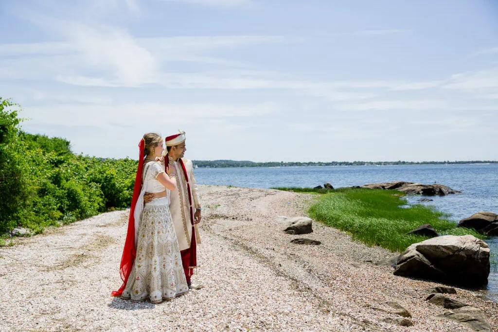 A bride and groom in indian wedding wear stand on a beach in rhode island looking out at the sea