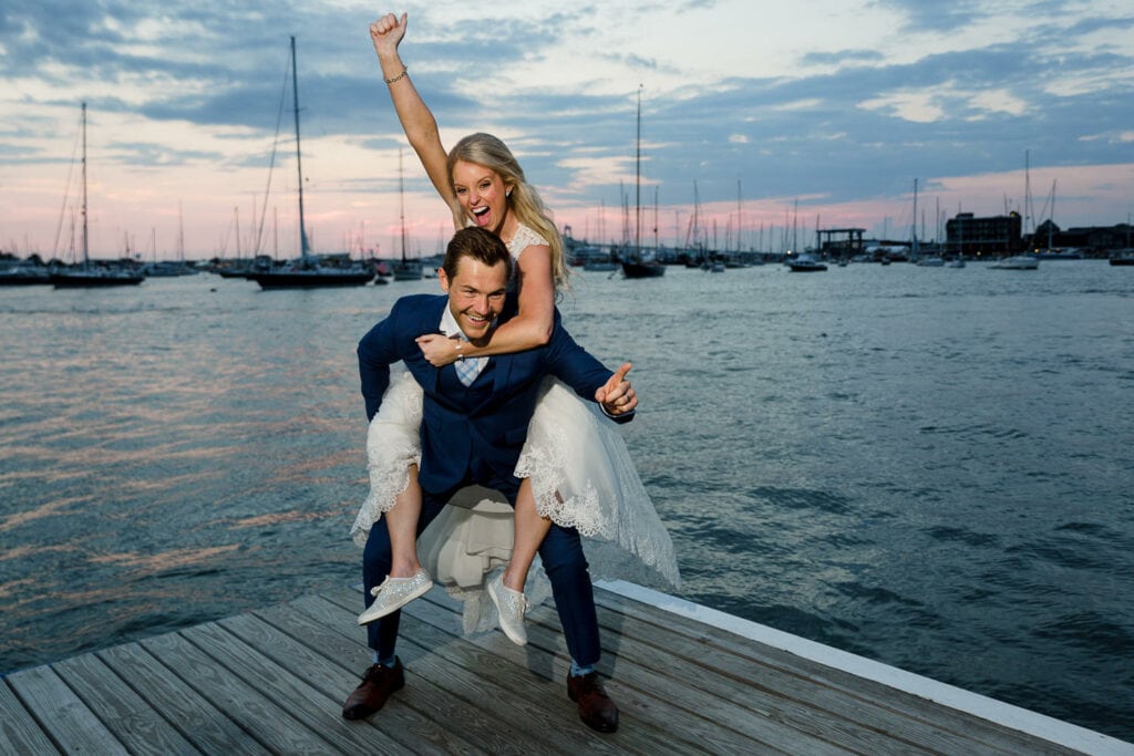 A bride riding piggyback on her groom with arm raised at the edge of a dock