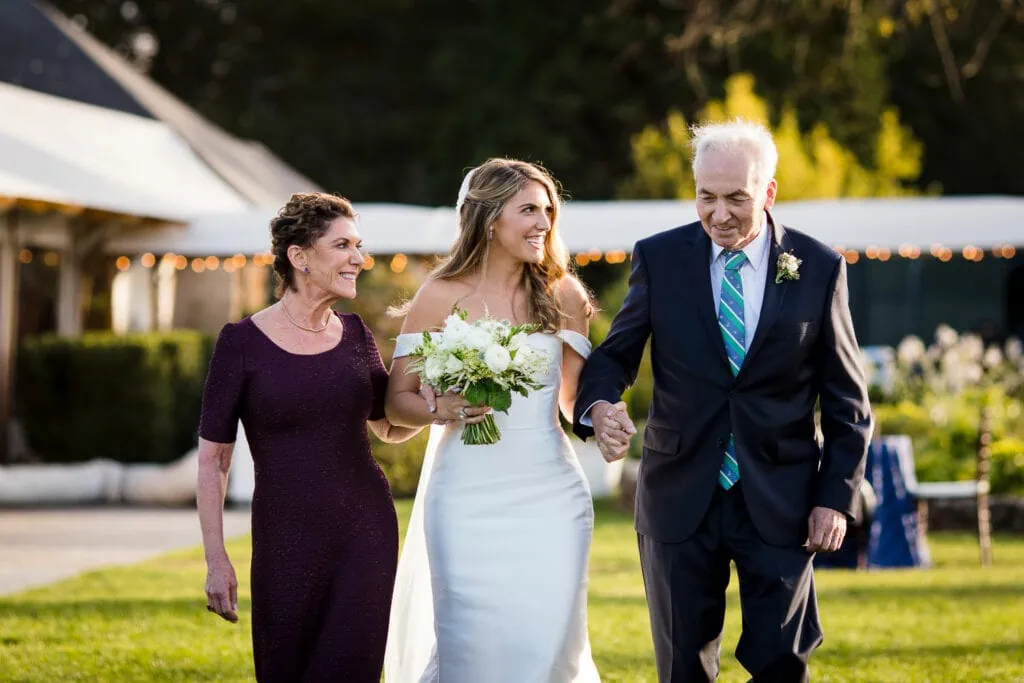 A bride walks down a grassy aisle with a woman and man on each arm