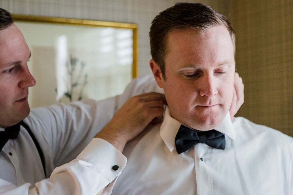 A groomsmen helping a groom adjust his collar and bowtie