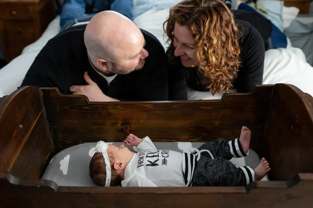 A mom and dad look at each other as their baby looks at them from her bassinet during an in-home newborn photo session