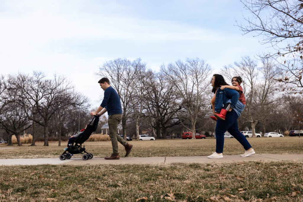 A dad pushing a stroller and mom carrying a little girl piggyback walk through a park