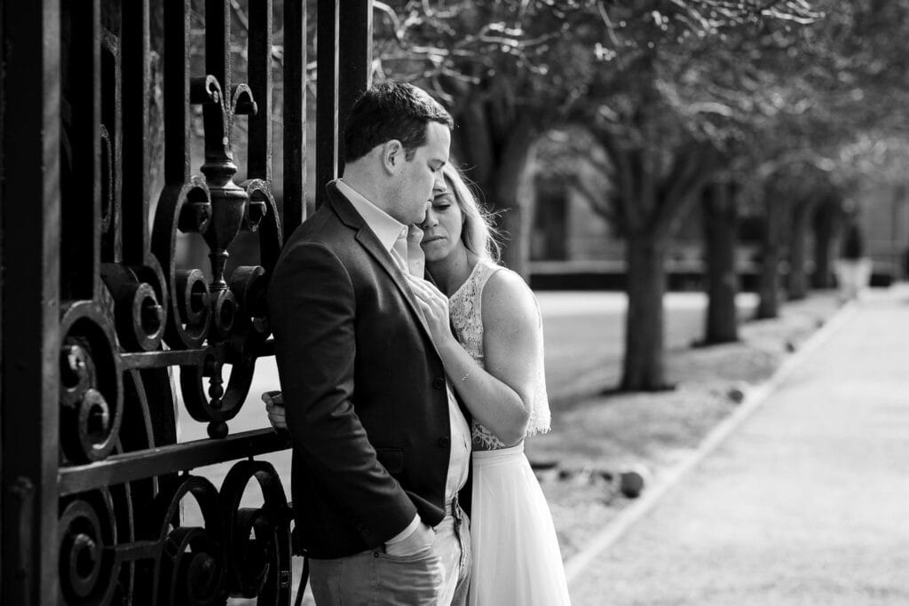 A woman snuggles against a mans shoulder as he leans on a wrought iron gate