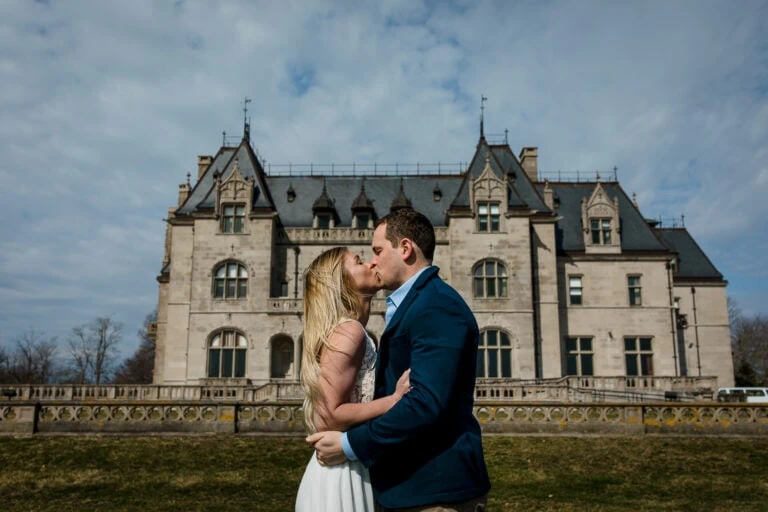 A woman in white and man in a navy suit kiss and embrace with ochre court mansion behind them