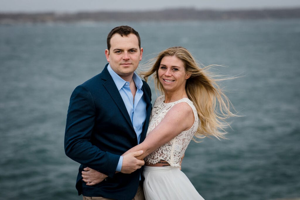 A man and woman embrace and look at the camera for a photo with the ocean in the background