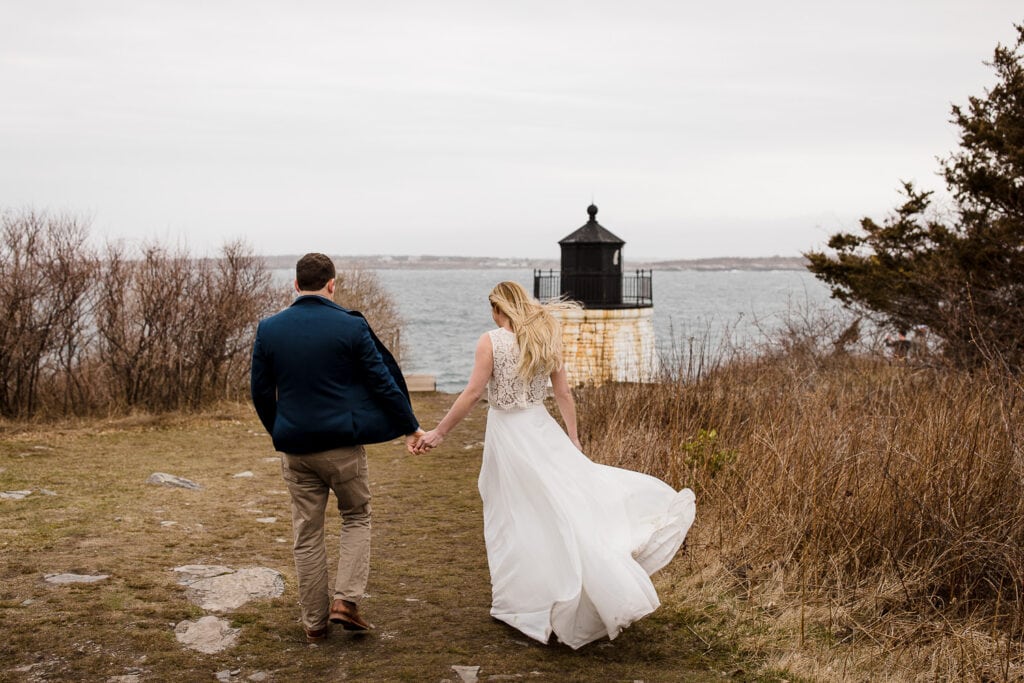 A man and woman walk toward a lighthouse and the ocean with their clothing blowing in the wind