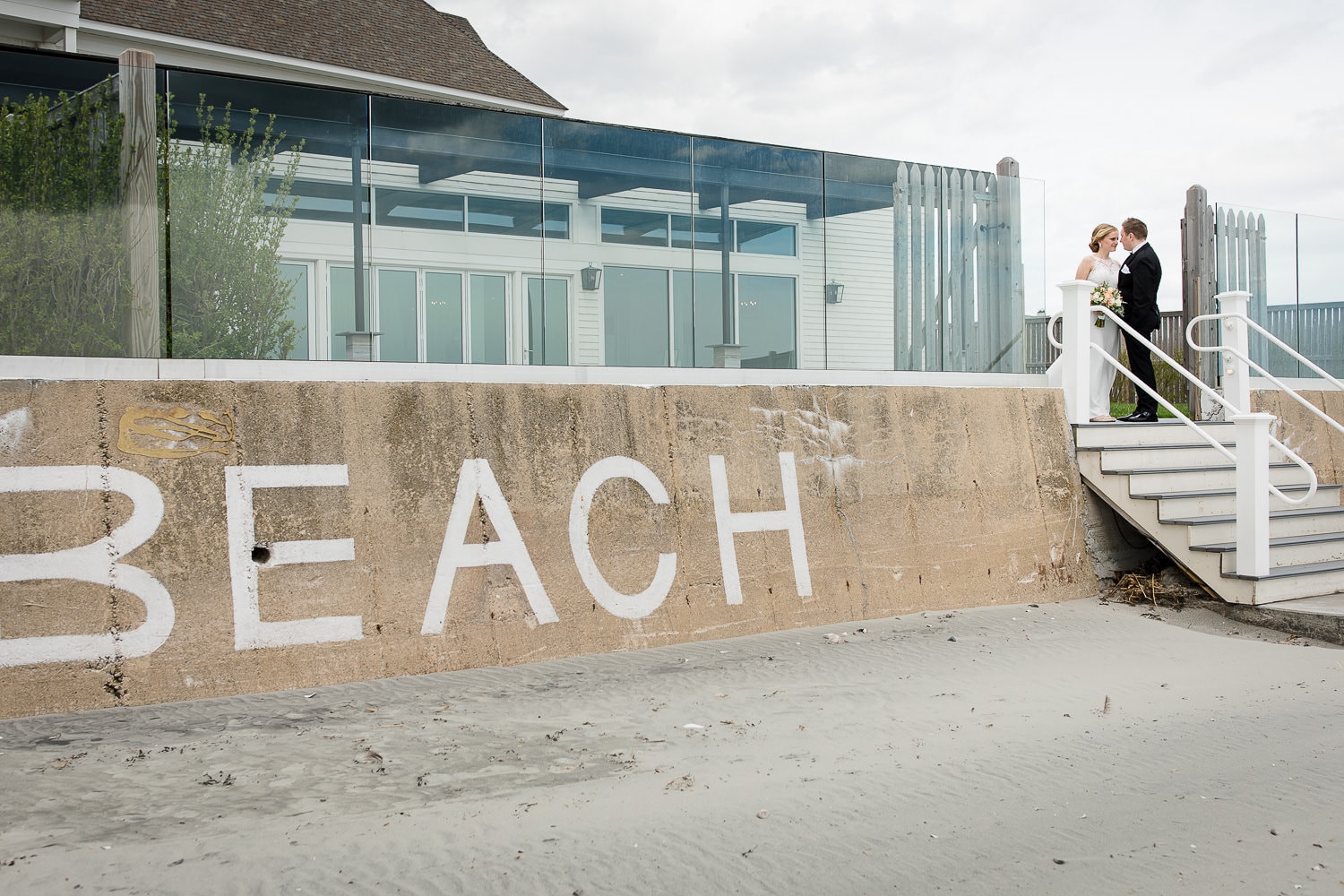A bride and groom stand at the top of a set of stairs leading to the beach with the word BEACH written on the sea wall in front of their wedding at the Newport Beach House in Middletown, RI