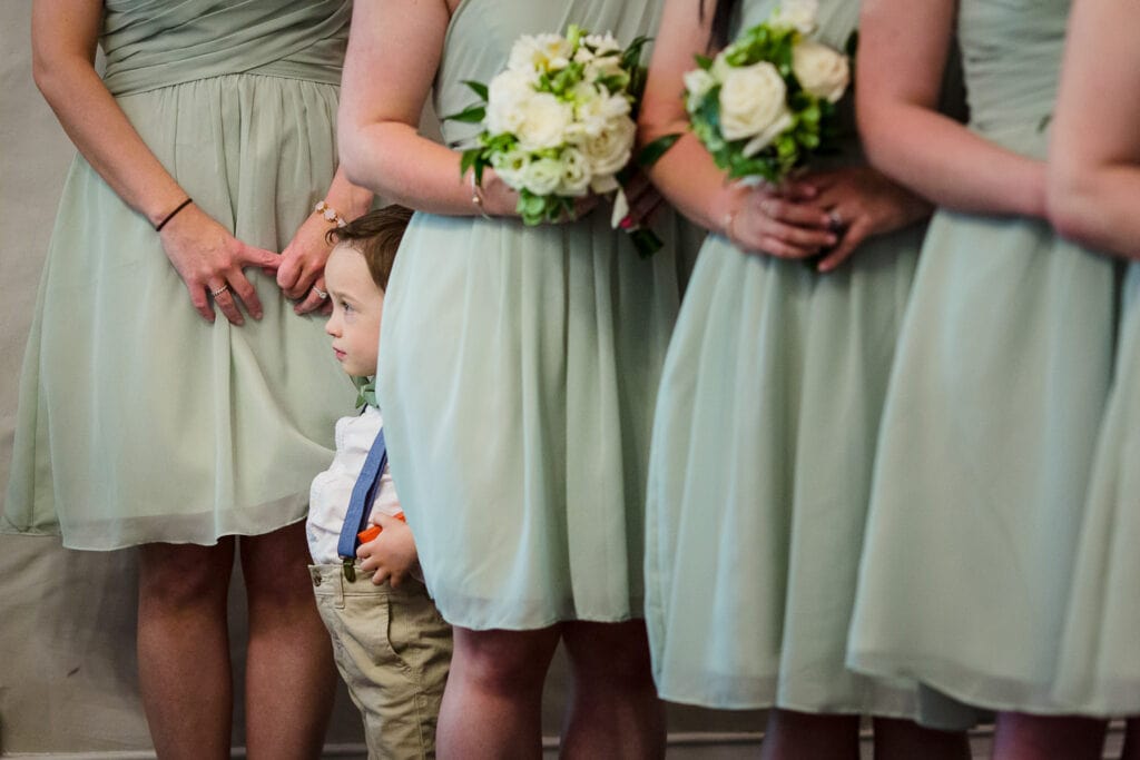 A ring bearer stands amidst the legs of a row of bridesmaids dressed in green