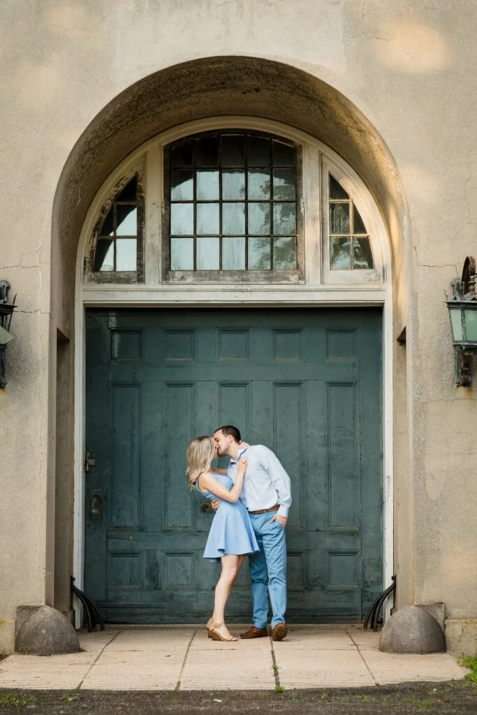 A man and woman wearing light blue kiss in front of the blue carriage house door for their engagement photosat harkness memorial state park