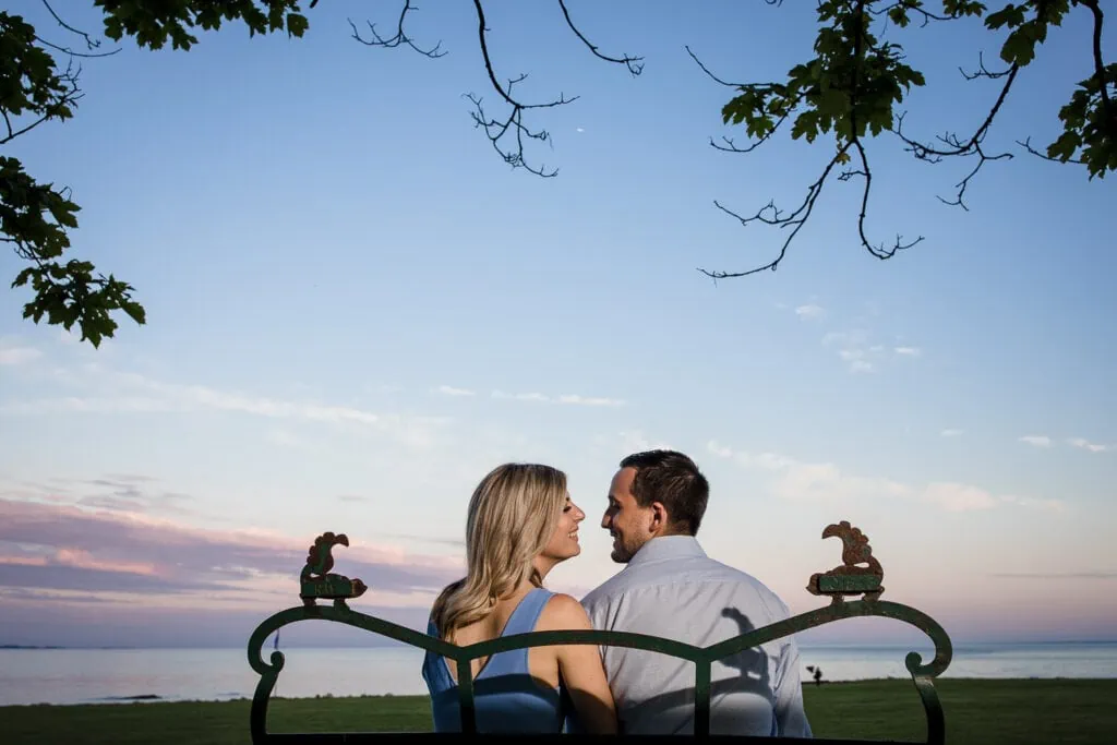 A man and woman sit looking at each other on a bench at sunset to end their harkness memorial state park engagement photos