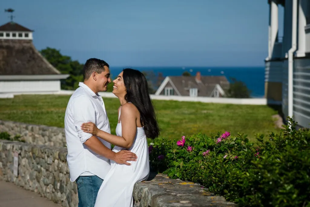 A woman sits on a stone wall with a man standing in front of her and a lawn, roses and the ocean behind them