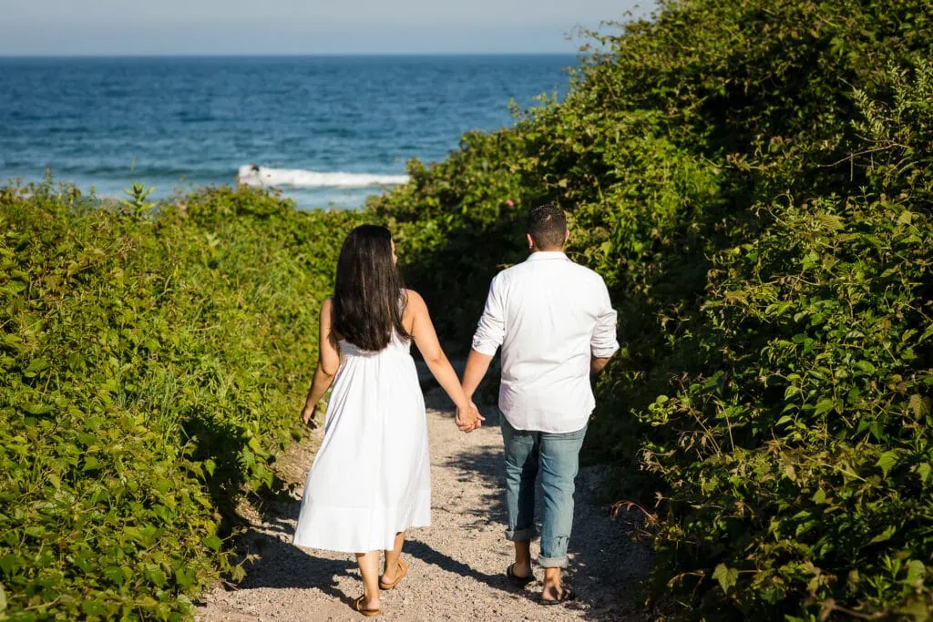 A man and woman in white walk down a rose lines pathway to the beach at their watch hill engagement photos