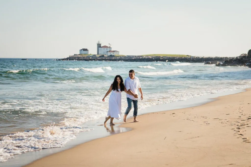 A woman in white and man in white laugh and run away from the surf holding hands at their watch hill engagement session