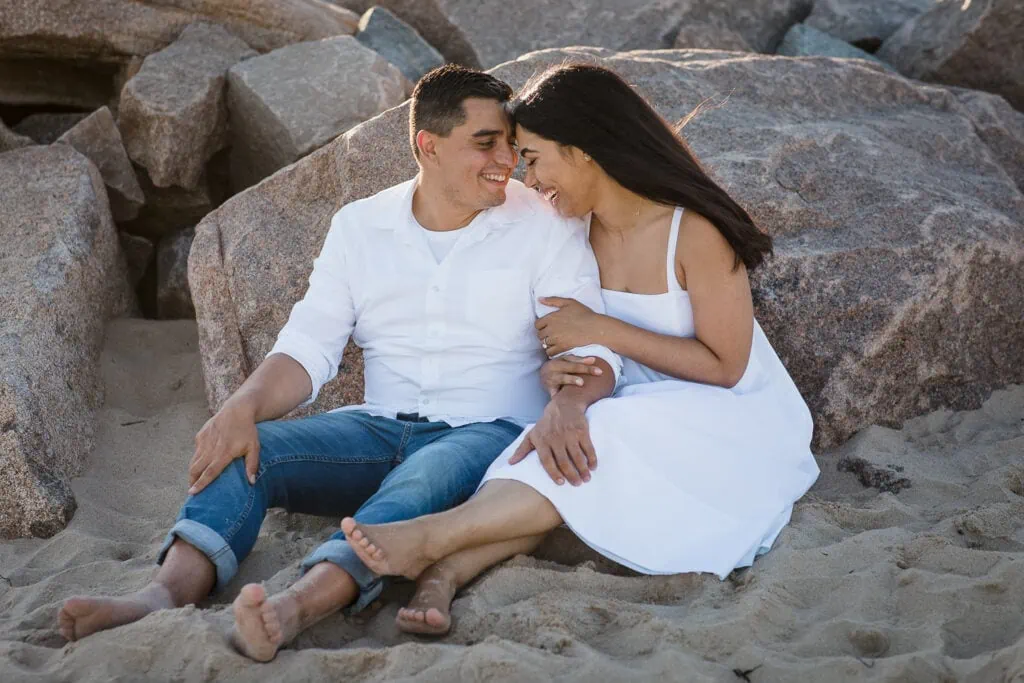 A man and woman wearing white sit in the sand and cuddle in front of rocks
