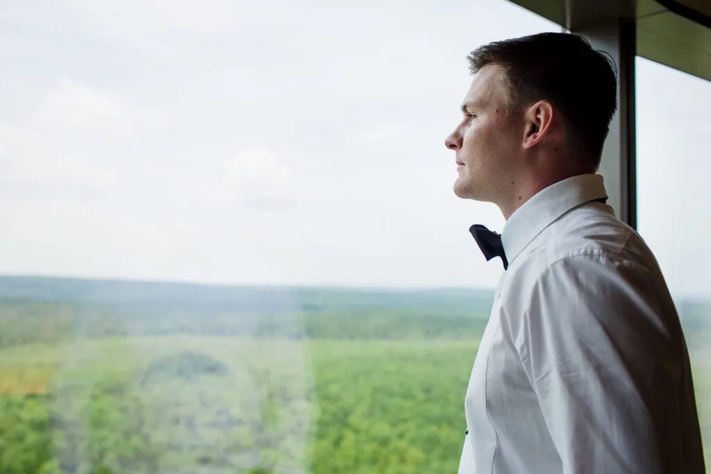 A man in white shirt and bowtie looks out a window at a lush green landscape