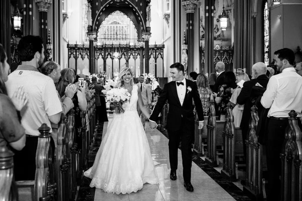 A bride and groom walk hand in hand out of their church wedding with guests clapping in the pews on either side