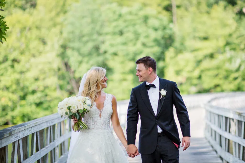 A blonde woman in white lace dress holding flowers walks hand in hand with a man wearing a black tuxedo and bowtie on a wooden bridge at their lake of isles wedding