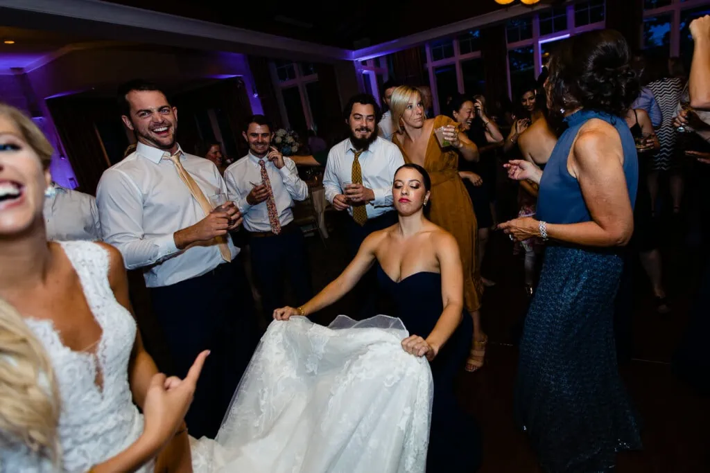 A bride laughs and points as her bridesmaid fans her with her dress train