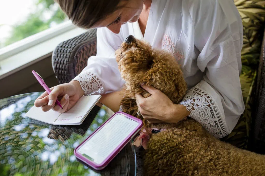 A woman in white robe writing in a notebook pets her goldendoodle dog