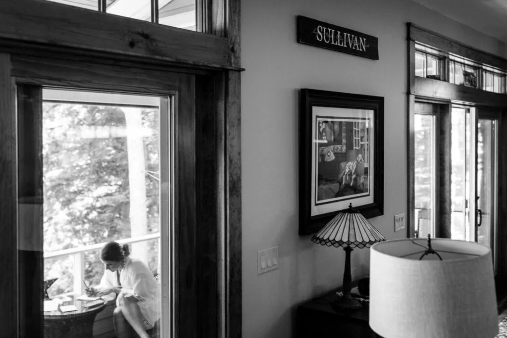 A woman out on a porch writing in a robe taken from inside a lake house with the name Sullivan on the wall