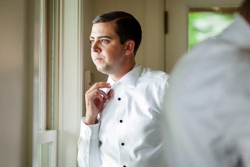 A man adjusts the collar of his white dress shirt as he looks out a window