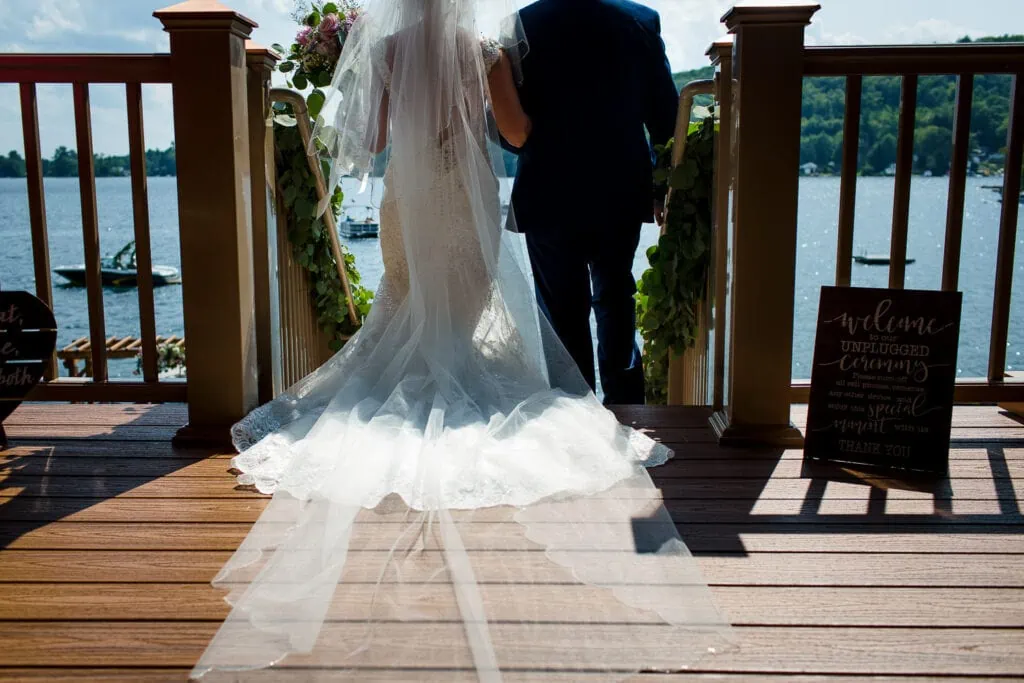 The back of a bride with long veil and main in navy walking down a set of deck stairs towards a lake