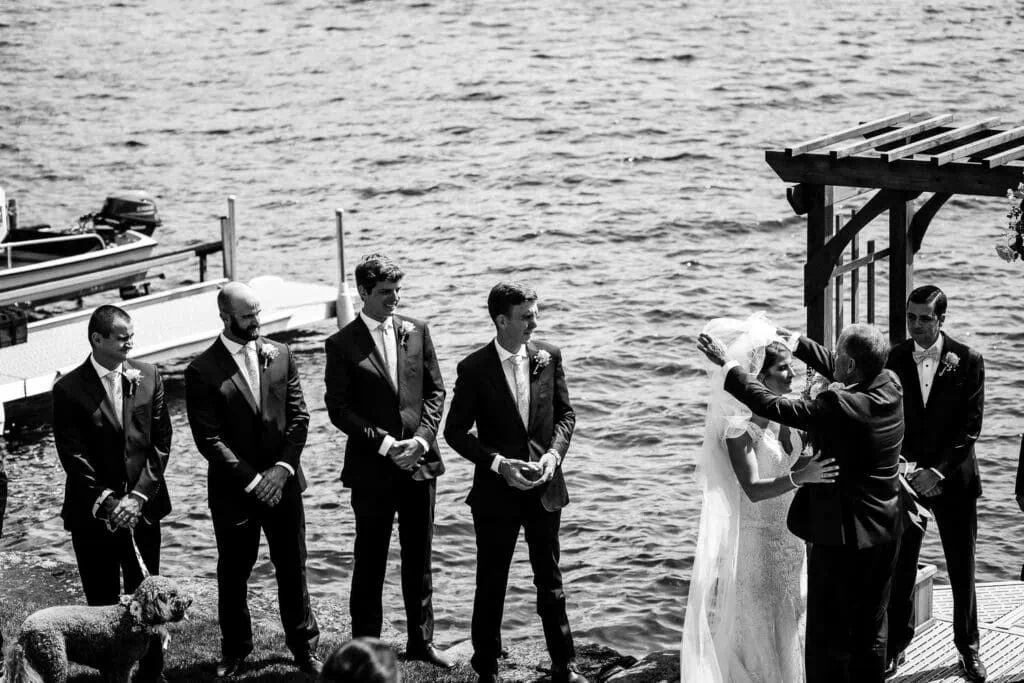 A father removes a veil from over his daughters face as a row of groomsmen look on at the edge of a lake