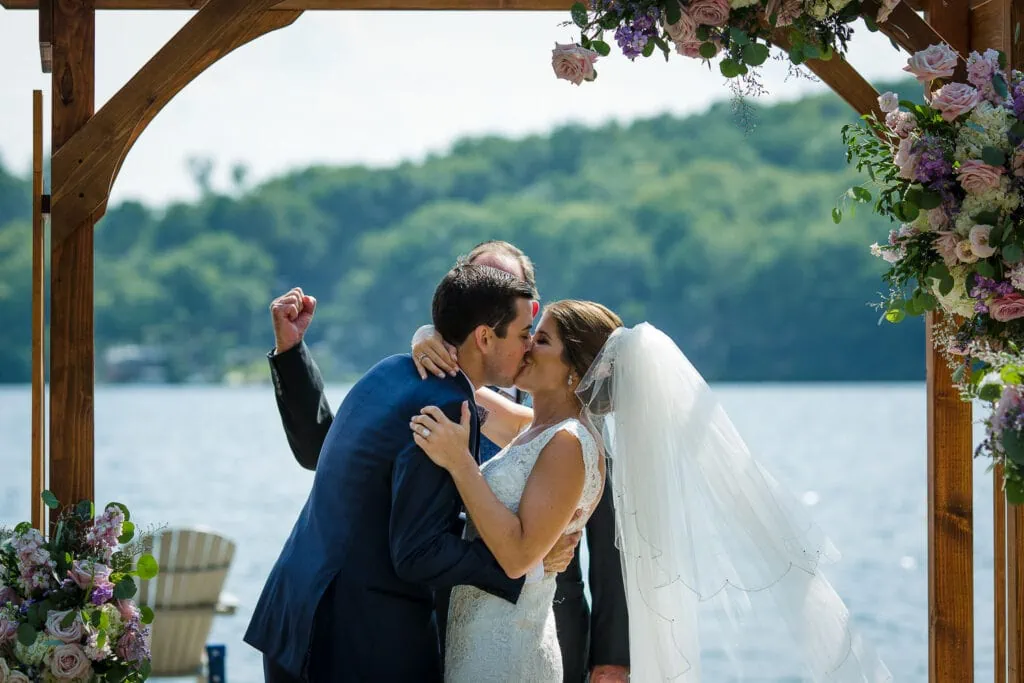 A hand fist pumps behind a couple sharing their first kiss at the end of their wedding ceremony by a lake