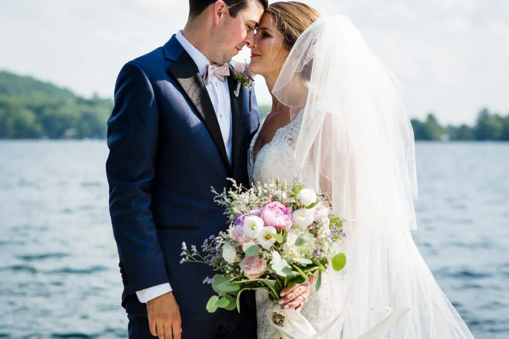 A bride and groom touch foreheads near a lake