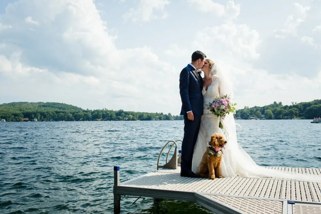 A bride and groom kiss on a dock by a lake with a goldendoodle dog in front of them
