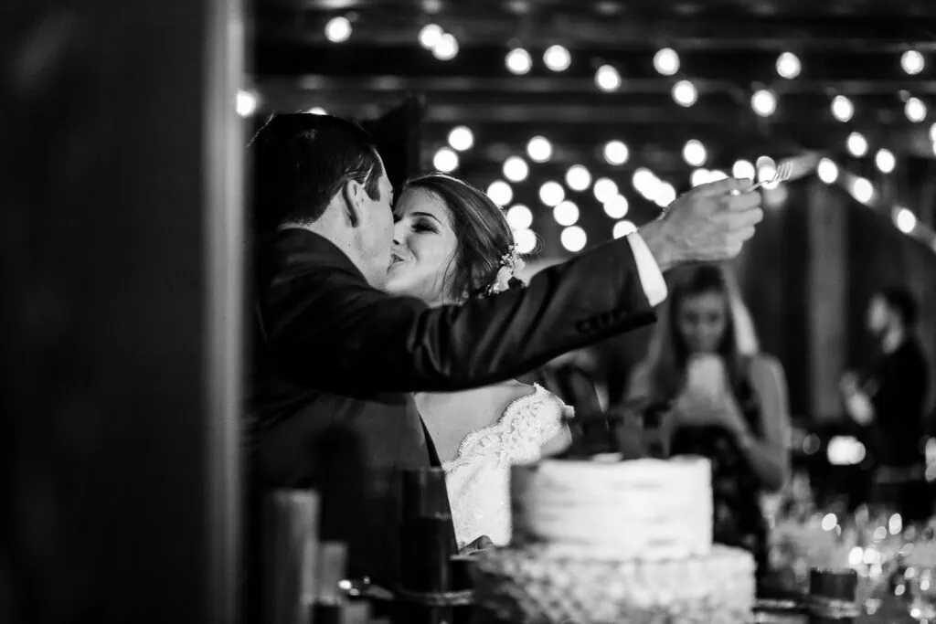 A bride kisses a groom who is holding a fork out above a wedding cake