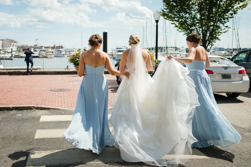 Two women in light blue dresses helps a bride walk across a crosswalk to where the groom is waiting with his back turned by a fence and the water