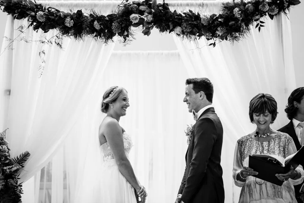 A bride and groom laughing at a ceremony in front of a sheer curtain and a flower garland