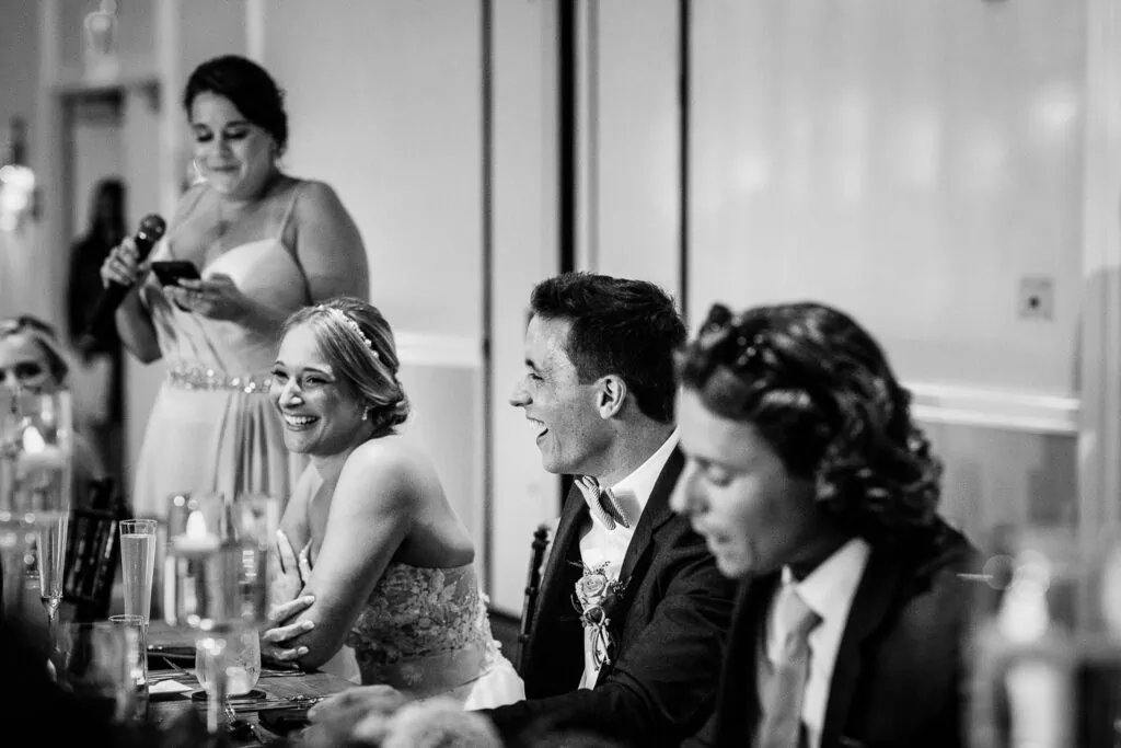 A bride and groom reacting to a woman giving a speech