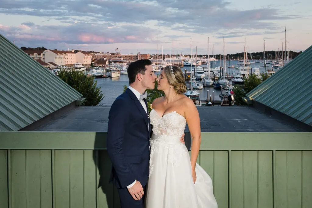 A bride and groom kissing on top of a building at sunset with a marina of boats in the distance