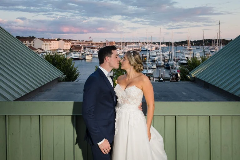 A bride and groom kissing on top of the Newport Marriott at sunset with a marina of boats in the distance on their wedding night