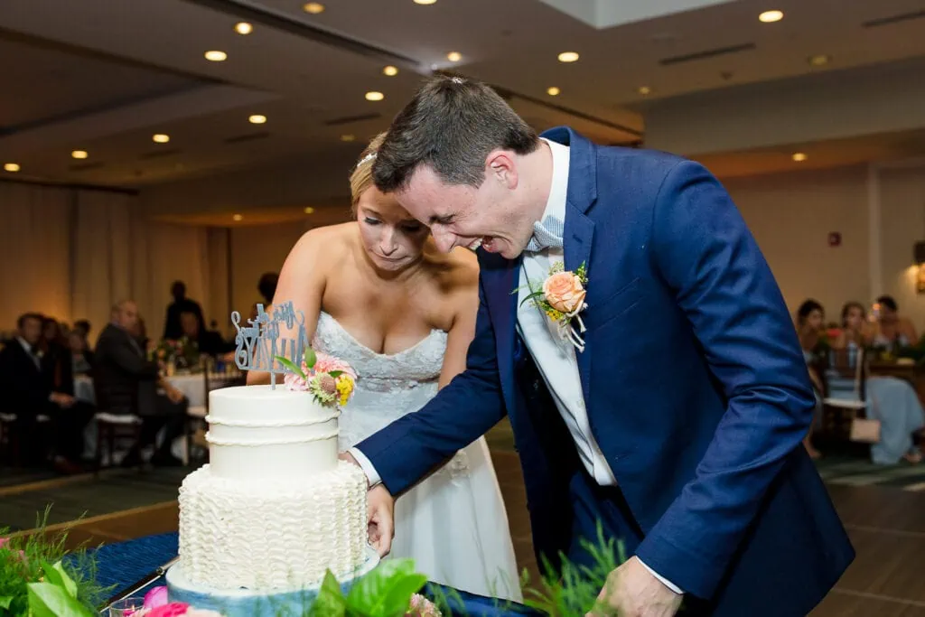 A groom laughing hysterically as the bride cringes as they cut their wedding cake