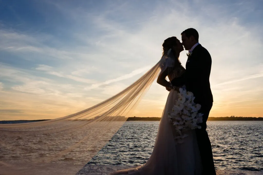 A bride and groom kissing in silhouette with a long flowing veil beneath a sunset sky