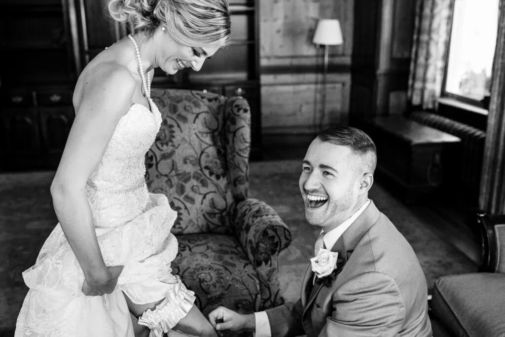 A bride laughs and holds up her dress skirt as a man also laughs while placing her garter on her leg