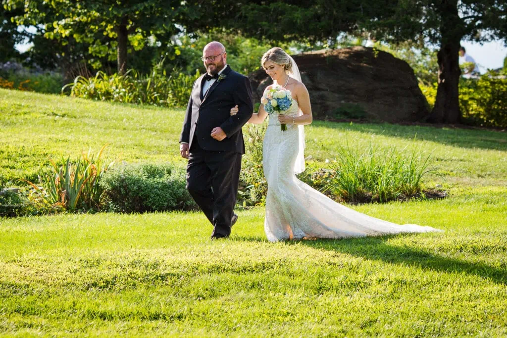 A bride in white with her father in arm walk down a grassy slope