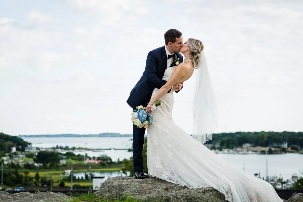 A groom dipping and kissing his bride atop a rock with the coastline in the distance at their Inn at Mystic Wedding