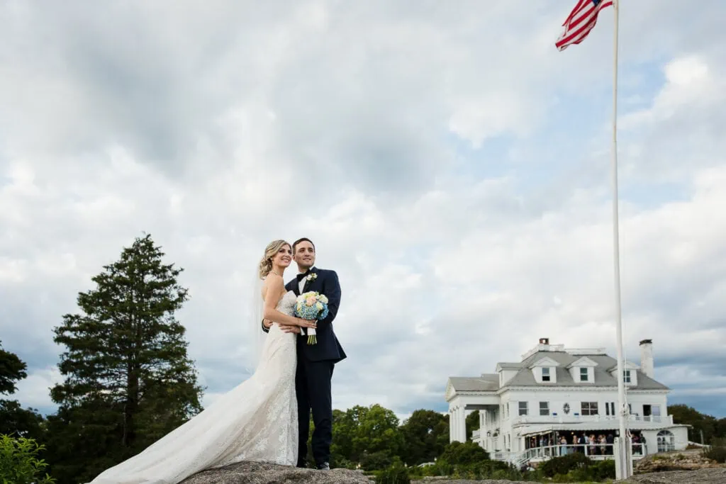 A bride and groom stand atop a rock looking off into the distance at their wedding with the Inn at Mystic in the background