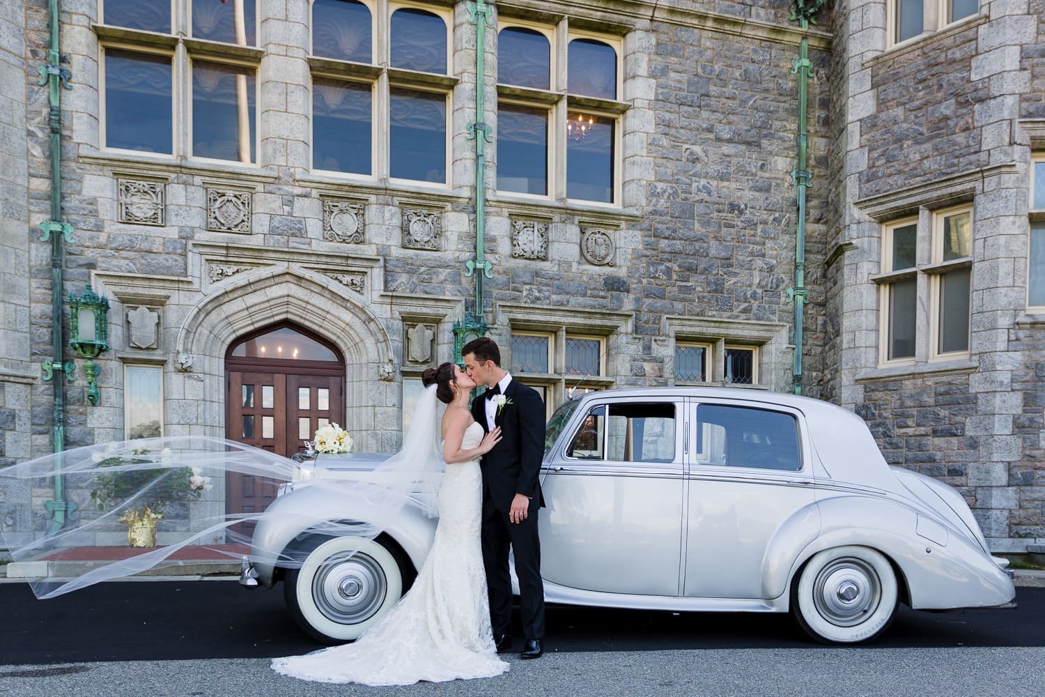 A bride and groom kiss in front of a vintage car and the branford house mansion